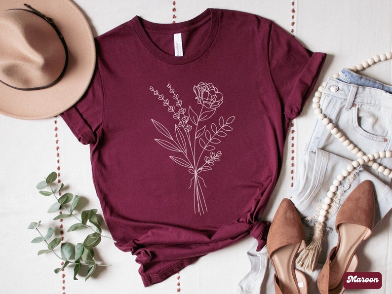 K&ouml;nnte beinhalten: Ein maroonfarbenes T-Shirt mit einer wei&szlig;en Linienzeichnung eines Blumenstrau&szlig;es. Die Blumen umfassen eine gro&szlig;e Pfingstrose und kleinere Lavendelzweige.
