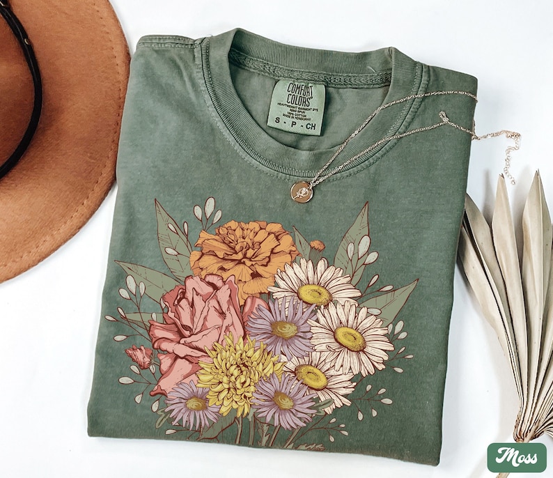 Pode incluir: Uma camiseta verde musgo com um design de buqu&ecirc; floral com flores laranja, rosa, amarelas e brancas. A camisa tem gola redonda e mangas curtas. Um colar de ouro com um pequeno pingente repousa sobre a camisa. A etiqueta diz "Comfort Colors".