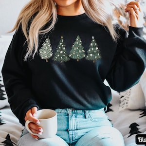 K&ouml;nnte beinhalten: Schwarzes Sweatshirt mit einem Motiv von vier Weihnachtsb&auml;umen, die mit Z&auml;hnen verziert sind. Die B&auml;ume sind gr&uuml;n mit wei&szlig;en Lichtern und einem goldenen Stern oben. Das Sweatshirt wird von einer Person getragen, die auf einem Sofa sitzt.