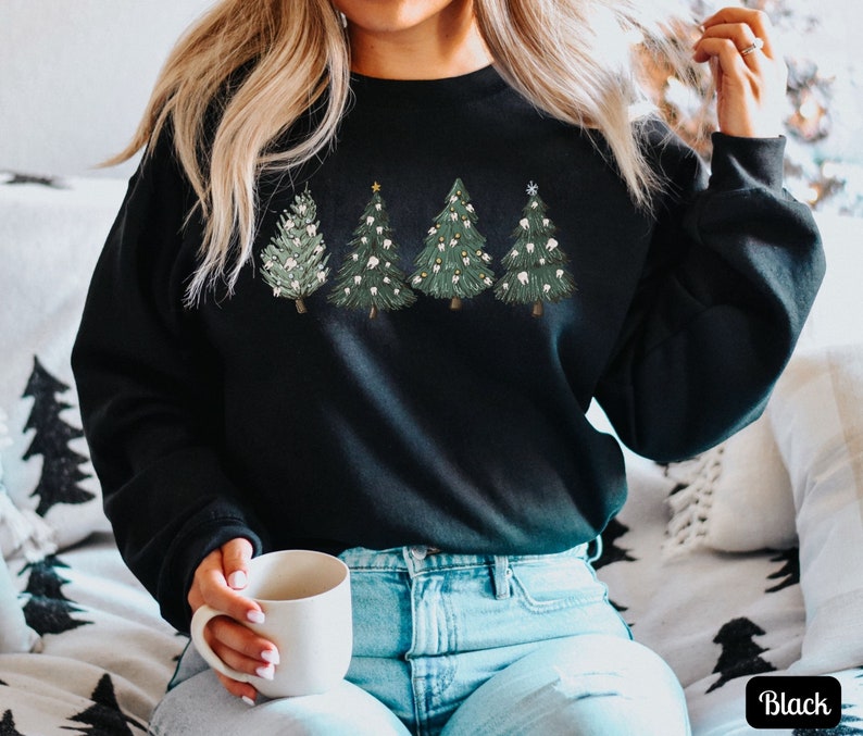 K&ouml;nnte beinhalten: Schwarzes Sweatshirt mit einem Motiv von vier Weihnachtsb&auml;umen, die mit Z&auml;hnen verziert sind. Die B&auml;ume sind gr&uuml;n mit wei&szlig;en Lichtern und einem goldenen Stern oben. Das Sweatshirt wird von einer Person getragen, die auf einem Sofa sitzt.