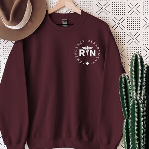 Peut inclure: Sweat-shirt marron avec un graphique blanc d'un symbole m&eacute;dical et le texte "Emergency Department RN".