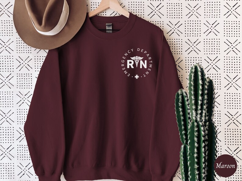 Peut inclure: Sweat-shirt marron avec un graphique blanc d'un symbole m&eacute;dical et le texte "Emergency Department RN".