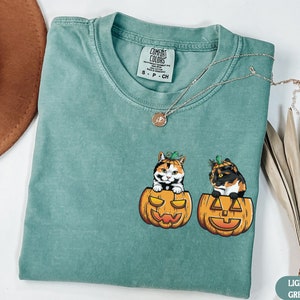 May include: A light green t-shirt with a graphic of two calico cats wearing pumpkin hats and sitting inside pumpkins. The text "COMFORT COLORS" is on a white tag attached to the shirt.
