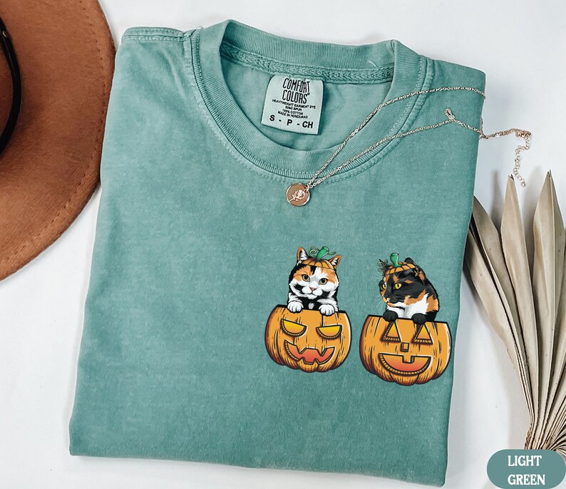 May include: A light green t-shirt with a graphic of two calico cats wearing pumpkin hats and sitting inside pumpkins. The text "COMFORT COLORS" is on a white tag attached to the shirt.