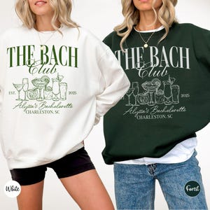 K&ouml;nnte beinhalten: Zwei wei&szlig;e und waldgr&uuml;ne Sweatshirts mit dem Text "The Bach Club" und einer Grafik von Cocktails. Der Text "Est. 2025" und "Alyssa's Bachelorette Charleston, SC" sind ebenfalls auf den Sweatshirts enthalten.
