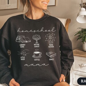 Puede incluir: Sudadera negra con texto blanco que dice "homeschool mama" e ilustraciones de libros, un &aacute;rbol, una persona leyendo, una taza de caf&eacute; y un libro con la palabra "grace" en &eacute;l. Las ilustraciones est&aacute;n dispuestas en una cuadr&iacute;cula.