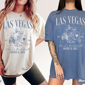 May include: Two t-shirts with a Las Vegas theme. The shirts are in ivory and blue jean colors. The design features a cocktail glass with dice and playing cards. The text on the shirts reads "Las Vegas" and "Olivia's Bachelorette Drink & Dice".