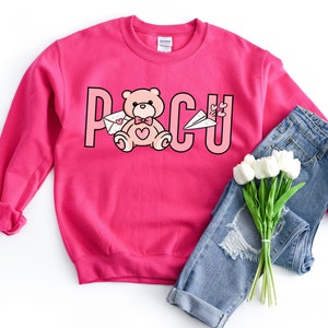Peut inclure: Sweat-shirt rose avec un motif repr&eacute;sentant un ours en peluche tenant une lettre et le texte "PCU". Le sweat-shirt est associ&eacute; &agrave; un jean bleu et un bouquet de fleurs blanches.