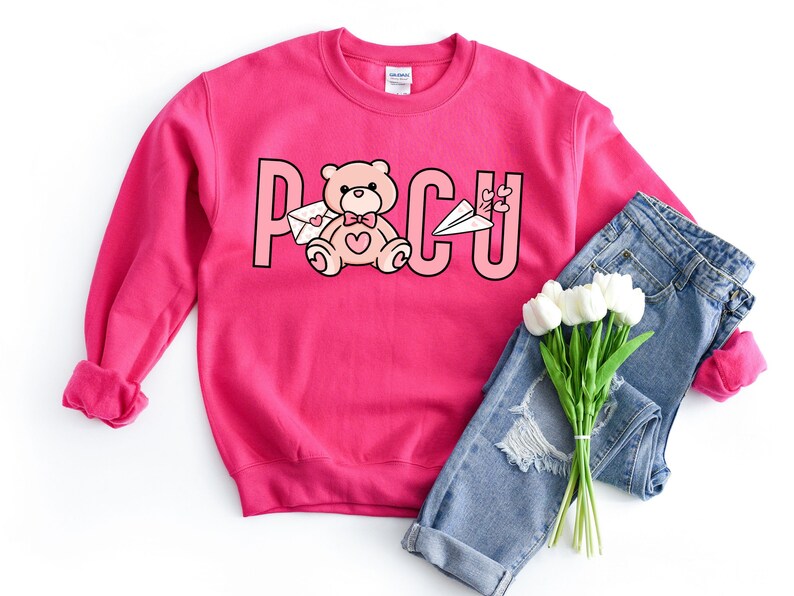 Peut inclure: Sweat-shirt rose avec un motif repr&eacute;sentant un ours en peluche tenant une lettre et le texte "PCU". Le sweat-shirt est associ&eacute; &agrave; un jean bleu et un bouquet de fleurs blanches.