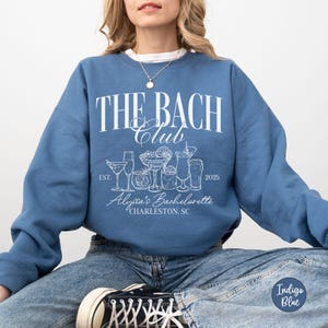 K&ouml;nnte beinhalten: Ein blaues Sweatshirt mit dem Text "The Bach Club" in Wei&szlig;, das eine Grafik von Cocktails und den Text "Alexa's Bachelorette Charleston, SC" in Wei&szlig; zeigt. Der Text "Est. 2025" ist ebenfalls in Wei&szlig;.