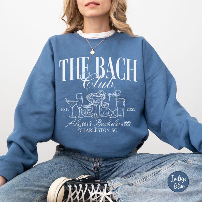 K&ouml;nnte beinhalten: Ein blaues Sweatshirt mit dem Text "The Bach Club" in Wei&szlig;, das eine Grafik von Cocktails und den Text "Alexa's Bachelorette Charleston, SC" in Wei&szlig; zeigt. Der Text "Est. 2025" ist ebenfalls in Wei&szlig;.