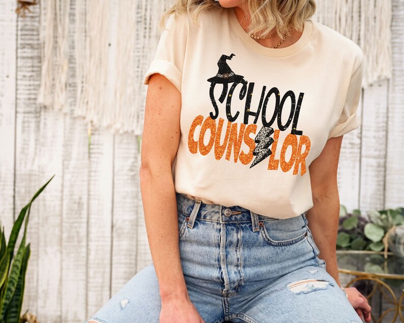 May include: A white t-shirt with the text "SCHOOL COUNSELOR" in orange and black glitter lettering. A black witch hat with a spider web design is above the text.