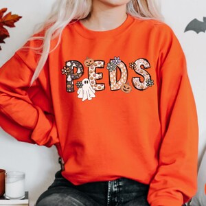 May include: An orange sweatshirt with the word "PEDS" in a decorative font. The letters are filled with Halloween-themed patterns, including pumpkins, ghosts, and flowers. The sweatshirt is a long-sleeved crew neck.