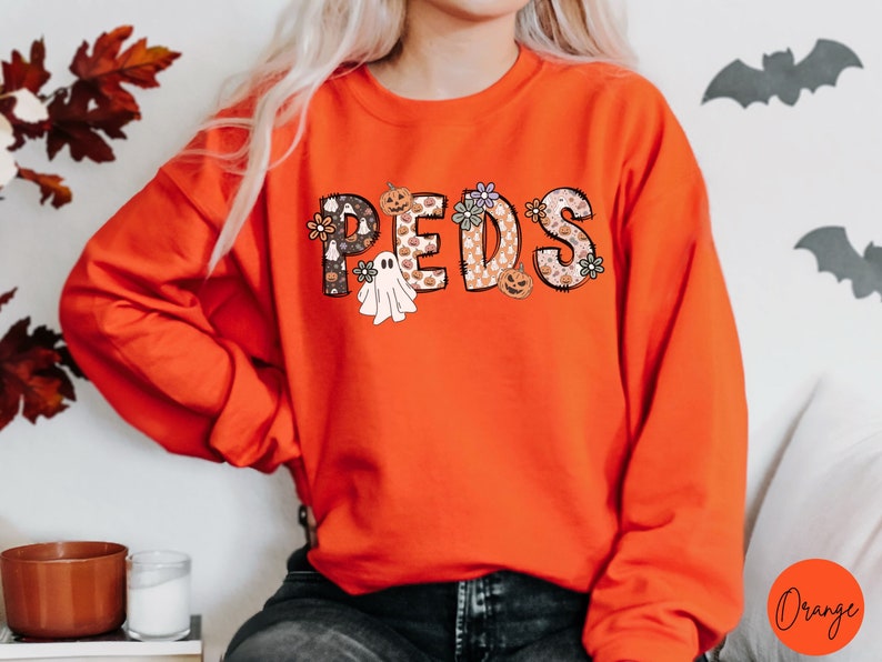 May include: An orange sweatshirt with the word "PEDS" in a decorative font. The letters are filled with Halloween-themed patterns, including pumpkins, ghosts, and flowers. The sweatshirt is a long-sleeved crew neck.