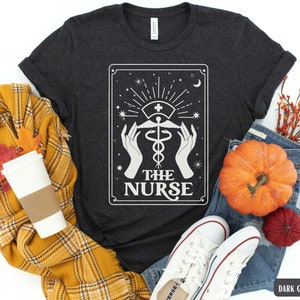 May include: Dark grey heather t-shirt with a white graphic of a tarot card design. The card features a caduceus symbol with the text "The Nurse".