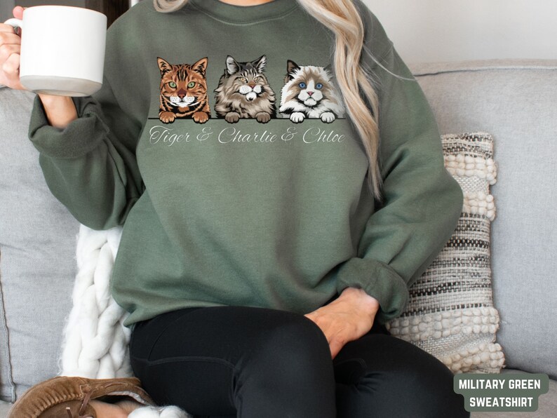Puede incluir: Una sudadera de color verde militar con un gr&aacute;fico de tres gatos asom&aacute;ndose por la parte superior. Los gatos se llaman Tiger, Charlie y Chloe.