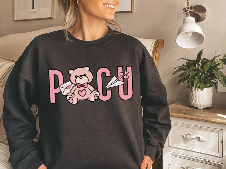 Peut inclure: Sweat-shirt noir avec un motif rose repr&eacute;sentant un ours en peluche tenant une lettre, un avion en papier et le mot "PCU".