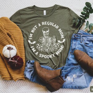 May include: A green t-shirt with the text "I'm not a regular mom, I'm a spooky mom!" and a skeleton graphic. The shirt is paired with blue jeans and brown ankle boots.