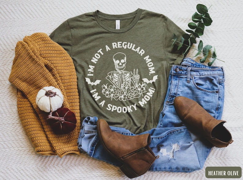 May include: A green t-shirt with the text "I'm not a regular mom, I'm a spooky mom!" and a skeleton graphic. The shirt is paired with blue jeans and brown ankle boots.