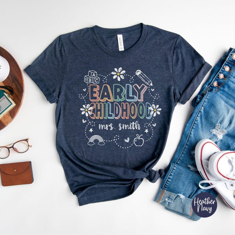 Pode incluir: Uma camiseta azul marinho com as palavras "EARLY CHILDHOOD Mrs. Smith" em letras coloridas, rodeada por ilustra&ccedil;&otilde;es l&uacute;dicas de um l&aacute;pis, flores, um arco-&iacute;ris e uma ma&ccedil;&atilde;. A camiseta &eacute; combinada com jeans azuis e t&ecirc;nis brancos.