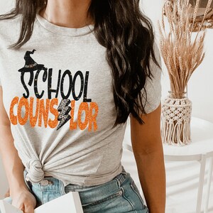 May include: A grey t-shirt with the text "SCHOOL COUNSELOR" in black and orange glitter. A black witch hat with an orange lightning bolt is above the word "SCHOOL".