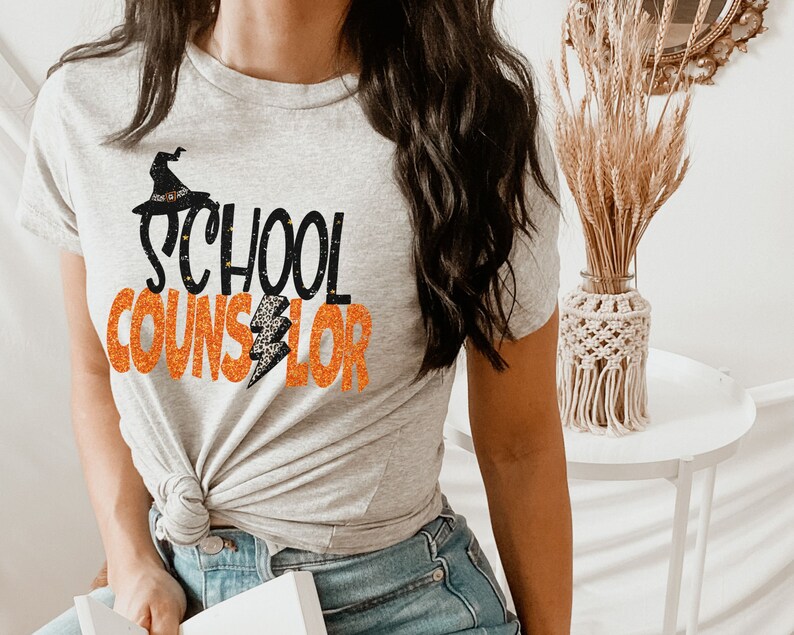 May include: A grey t-shirt with the text "SCHOOL COUNSELOR" in black and orange glitter. A black witch hat with an orange lightning bolt is above the word "SCHOOL".