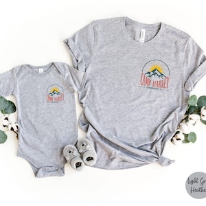 K&ouml;nnte beinhalten: Ein hellgrau meliertes T-Shirt und ein passender Baby-Body, beide mit einer kreisf&ouml;rmigen Grafik auf der Brust, die eine gelbe Sonne, blaue Berge und den roten Schriftzug "CAMP HARVEY" sowie darunter "PENNSYLVANIA, PA" in kleiner schwarzer Schrift zeigt.