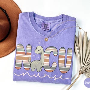 May include: Purple short-sleeved t-shirt with a striped design and a cartoon dinosaur. The shirt says "NCU nurse" in a pink font.