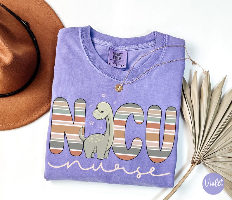 May include: Purple short-sleeved t-shirt with a striped design and a cartoon dinosaur. The shirt says "NCU nurse" in a pink font.