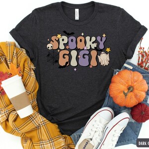 May include: Dark grey heather t-shirt featuring a retro-style graphic that says "Spooky Gigi" in colourful letters. The graphic also includes a ghost, a spider web, and bats.