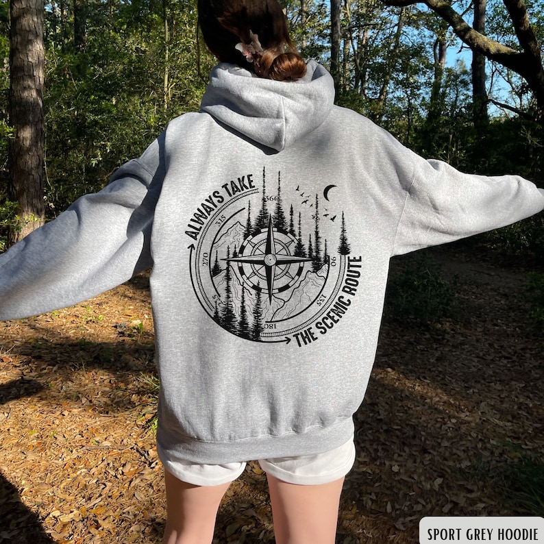 Może przedstawiać: Szara sportowa bluza z kapturem z czarnym wzorem kompasu na plecach. Wz&oacute;r kompasu przedstawia pasmo g&oacute;rskie, drzewa i napis "Always Take The Scenic Route".