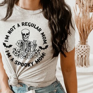 May include: A gray t-shirt with a black graphic of a skeleton holding a peace sign. The graphic is surrounded by flowers and bats. The text on the shirt reads "I'm not a regular mom. I'm a spooky mom!"