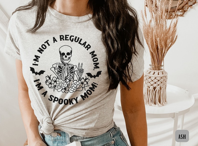 May include: A gray t-shirt with a black graphic of a skeleton holding a peace sign. The graphic is surrounded by flowers and bats. The text on the shirt reads "I'm not a regular mom. I'm a spooky mom!"