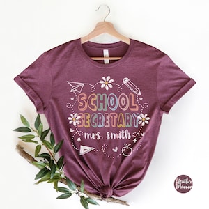 Peut inclure: T-shirt bordeaux avec l'inscription "SCHOOL SECRETARY" en lettres color&eacute;es, ainsi qu'un crayon, un avion en papier, des fleurs et une pomme. Le texte "mrs. smith" est &eacute;galement inclus. Le t-shirt est suspendu &agrave; un cintre en bois.