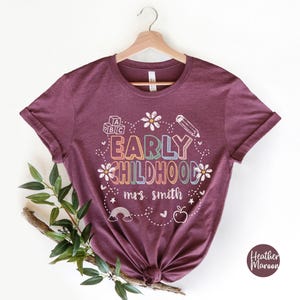 Pode incluir: Uma t-shirt bord&ocirc; com as palavras "EARLY CHILDHOOD" em letras coloridas, rodeada por um l&aacute;pis, flores, um arco-&iacute;ris e uma ma&ccedil;&atilde;. O texto "mrs. smith" est&aacute; abaixo. A t-shirt est&aacute; pendurada num cabide de madeira.