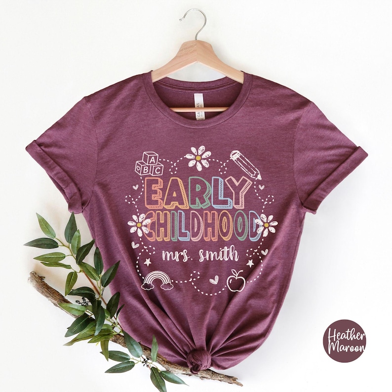 Pode incluir: Uma t-shirt bord&ocirc; com as palavras "EARLY CHILDHOOD" em letras coloridas, rodeada por um l&aacute;pis, flores, um arco-&iacute;ris e uma ma&ccedil;&atilde;. O texto "mrs. smith" est&aacute; abaixo. A t-shirt est&aacute; pendurada num cabide de madeira.