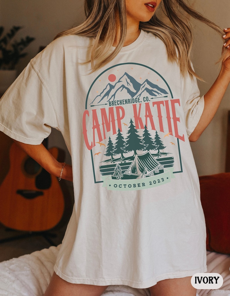 May include: Ivory oversized t-shirt with a graphic print of a mountain scene with a tent and the text "Camp Katie - Breckenridge, Co. - October 2023."
