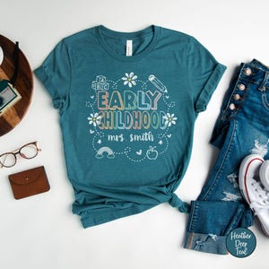 Pode incluir: T-shirt azul-petr&oacute;leo com as palavras "EARLY CHILDHOOD" em letras coloridas, rodeadas por um desenho floral, um l&aacute;pis, um arco-&iacute;ris e uma ma&ccedil;&atilde;. O texto "mrs. smith" est&aacute; abaixo. A camisa &eacute; combinada com jeans azuis e t&ecirc;nis brancos.