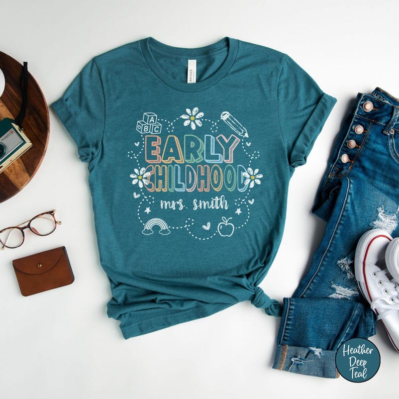 Pode incluir: T-shirt azul-petr&oacute;leo com as palavras "EARLY CHILDHOOD" em letras coloridas, rodeadas por um desenho floral, um l&aacute;pis, um arco-&iacute;ris e uma ma&ccedil;&atilde;. O texto "mrs. smith" est&aacute; abaixo. A camisa &eacute; combinada com jeans azuis e t&ecirc;nis brancos.