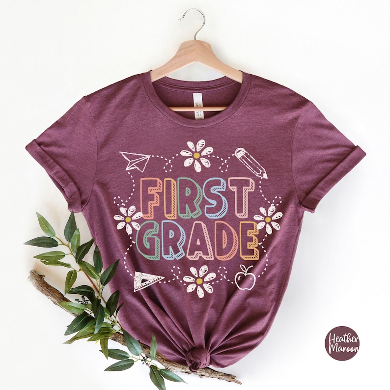 May include: A maroon t-shirt with the words "FIRST GRADE" in colorful letters, surrounded by white daisies, a paper airplane, a pencil, a triangle, and an apple. The shirt is hanging on a wooden hanger.