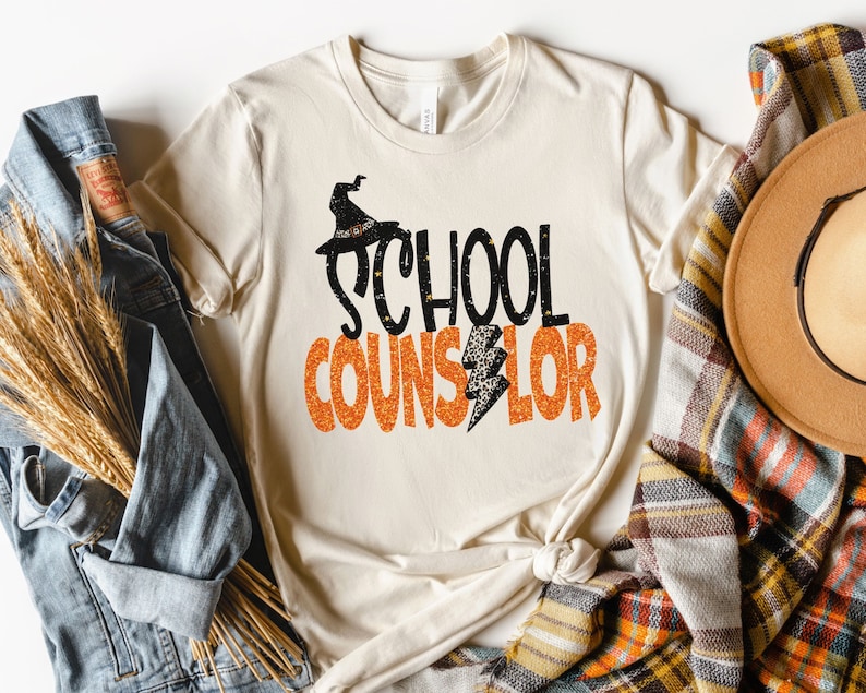May include: A beige t-shirt with a black witch hat and the text "SCHOOL COUNSELOR" in orange glitter.