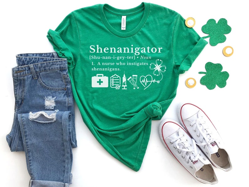 May include: Green t-shirt with white text that reads "Shenanigator [Shu-nan-i-gey-ter]. Noun 1. A nurse who instigates shenanigans."  The shirt has a graphic of a first aid kit, a clipboard, a shot glass, a heart with a stethoscope, and a four-leaf clover.