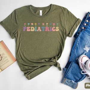 Puede incluir: Una camiseta verde con la palabra "PEDIATRICS" en letras arco&iacute;ris y una fila de flores coloridas sobre el texto. La camiseta est&aacute; sobre un fondo blanco con un par de pantalones vaqueros azules y zapatillas blancas.