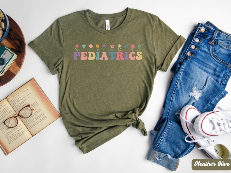 Puede incluir: Una camiseta verde con la palabra "PEDIATRICS" en letras arco&iacute;ris y una fila de flores coloridas sobre el texto. La camiseta est&aacute; sobre un fondo blanco con un par de pantalones vaqueros azules y zapatillas blancas.