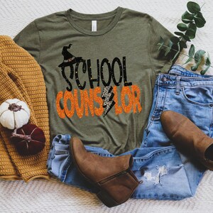 May include: A green t-shirt with the text "School Counselor" in black and orange glitter lettering. A witch's hat with a black and white pattern is above the text. The shirt is lying on a white surface with a brown sweater, blue jeans, and brown boots.