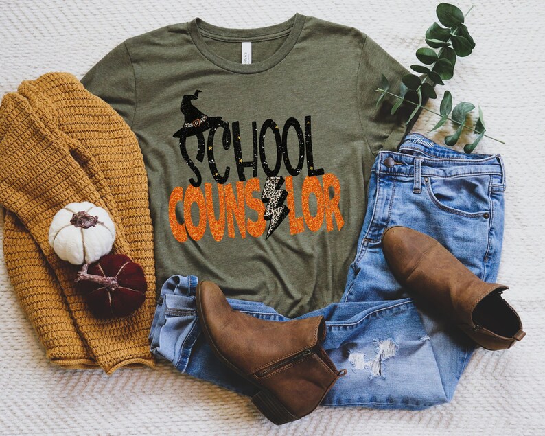 May include: A green t-shirt with the text "School Counselor" in black and orange glitter lettering. A witch's hat with a black and white pattern is above the text. The shirt is lying on a white surface with a brown sweater, blue jeans, and brown boots.