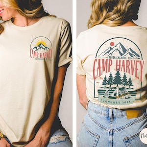 K&ouml;nnte beinhalten: Ein cremefarbenes Kurzarm-T-Shirt mit einem Grafikdesign. Auf der Brust befindet sich ein Berg- und Sonnenlogo mit dem Text Camp Harvey Fredericksburg, TX. Die R&uuml;ckseite zeigt eine gr&ouml;&szlig;ere Version derselben Berg-, Wald- und Zelt-Szene.