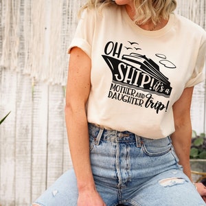 May include: A woman wearing a cream coloured t-shirt with a black graphic of a cruise ship and the text "Oh Ship! It's a Mother and Daughter Trip!" The shirt is a relaxed fit and has rolled up sleeves. The woman is wearing blue jeans with distressed details.