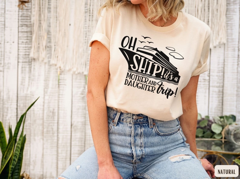 May include: A woman wearing a cream coloured t-shirt with a black graphic of a cruise ship and the text "Oh Ship! It's a Mother and Daughter Trip!" The shirt is a relaxed fit and has rolled up sleeves. The woman is wearing blue jeans with distressed details.