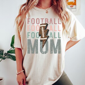 Op de afbeelding: Een cr&egrave;mekleurig T-shirt met een verweerde grafische afbeelding waarop "Football Mom" staat met een voetbal- en bliksemontwerp. De tekst is in tinten roze, groen en grijs.